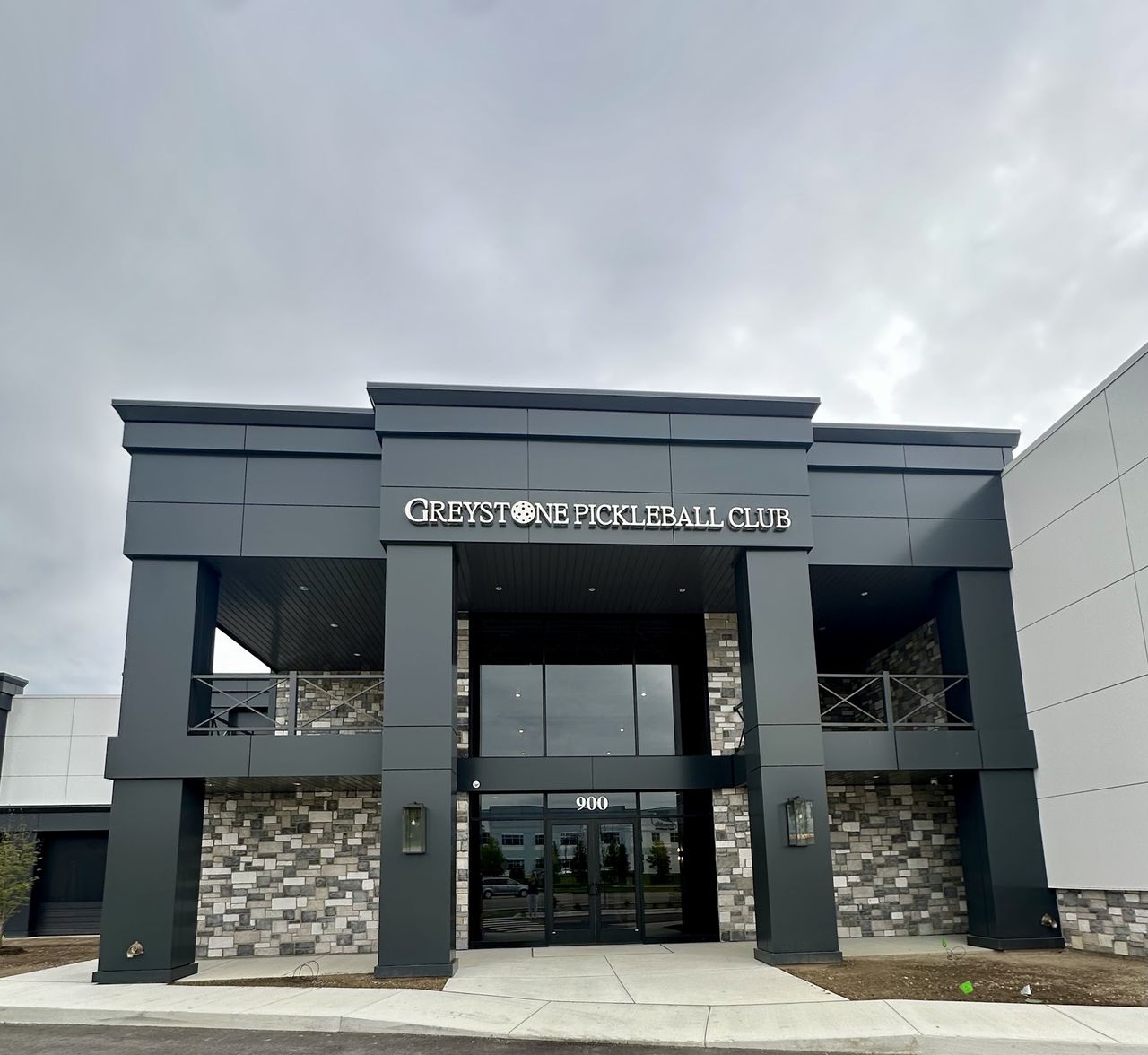 Exterior of Greystone Pickleball Club at 900 N Opdyke Rd in Auburn Hills, a dark two-story building with stone columns, glass doors, and the club sign above the entry
