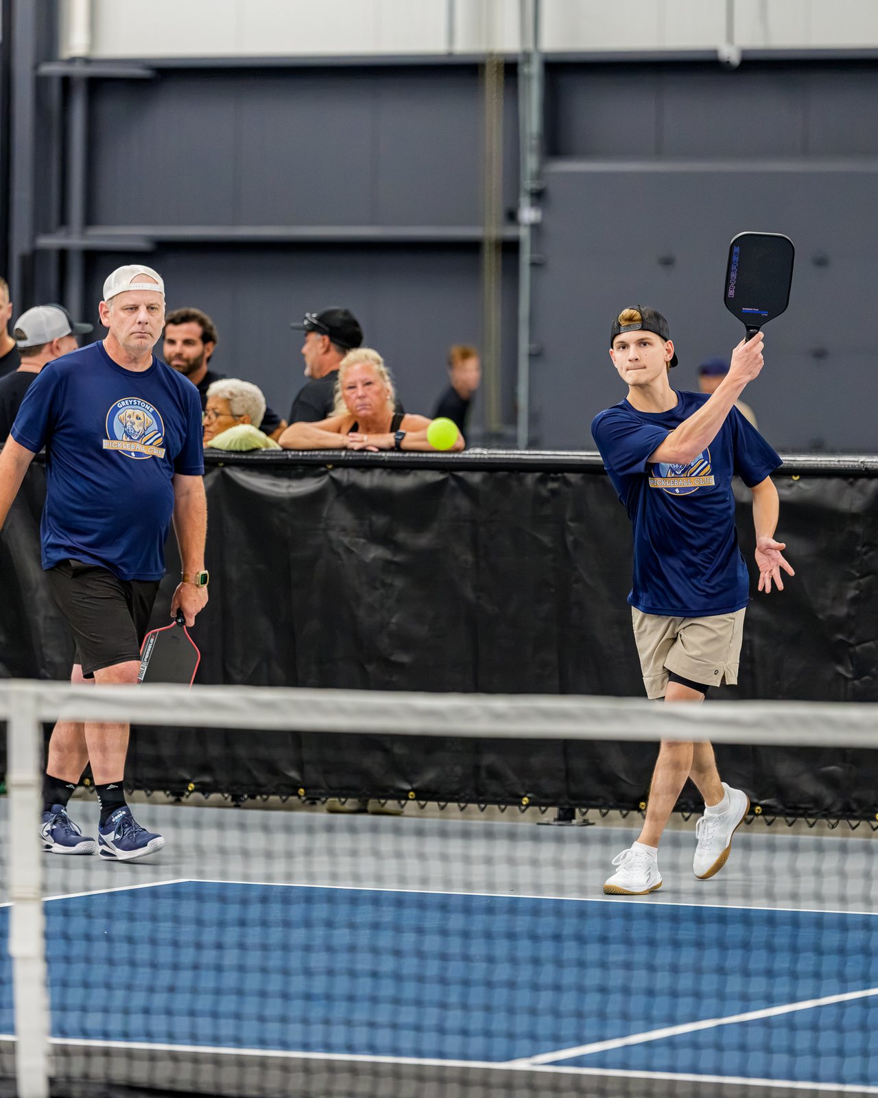 A Greystone league match: a player in a club shirt follows through at the net while spectators watch from the rail