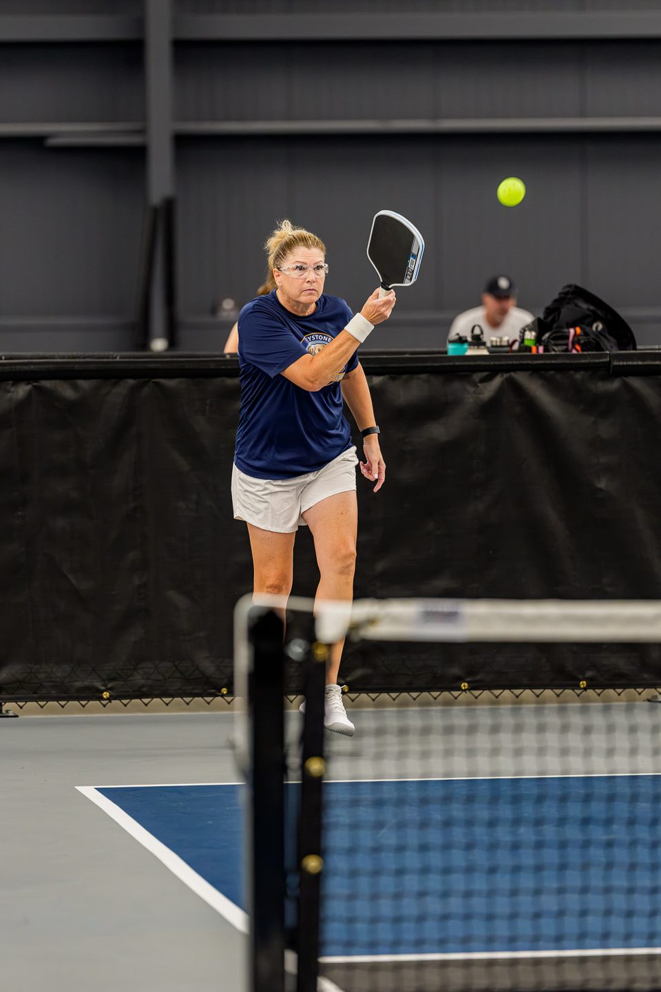 A Greystone coach in a club shirt lines up a forehand at the net on an indoor court