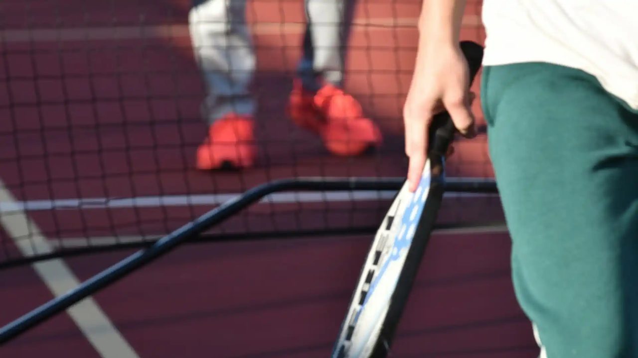 Close-up of a player gripping a paddle at the net on an indoor court at Greystone, opponents waiting on the far side