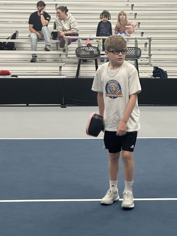 A young player holding a paddle on Stadium Court, with family watching from the bleachers